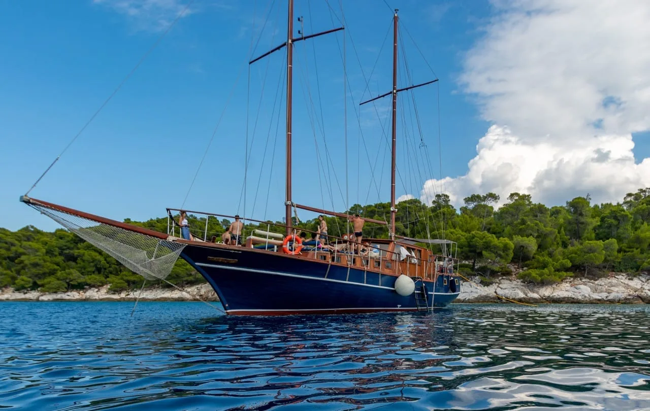 The blue-hulled two-masted wooden boat at anchor off a wooded shore