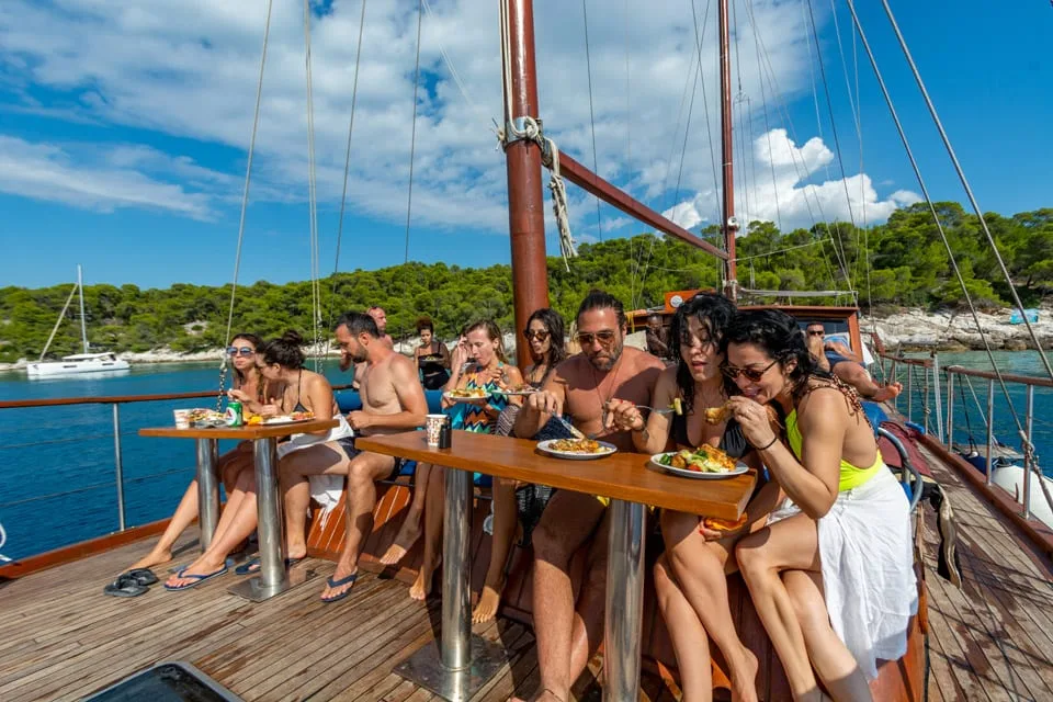 Guests eating lunch at deck tables with the boat anchored off a pine-covered shore
